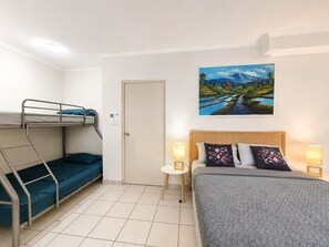 Ein Zimmer mit einem Etagenbett, einem Einzelbett, einer Tür, einem Wandbild und einem kleinen Tisch mit einer Lampe.