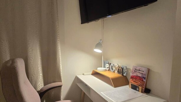 Una stanza con una scrivania bianca, una sedia, una TV a schermo piatto montata a parete, una lampada da scrivania e un libro intitolato 'Northern Territory'.