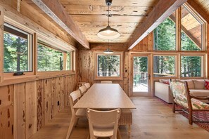 Interior de madera con una larga mesa de comedor, sillas y grandes ventanales que ofrecen vistas a un bosque.