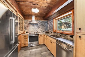 A kitchen with wooden cabinets, a stainless steel refrigerator, a built-in oven, and a window with a view of a wooden structure outside. - Marigold Cabin by Tahoe Getaways - 3BR in Alpine Meadows w/ Ski Shuttle! (Alpine Meadows)
