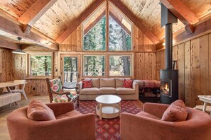A cozy cabin interior with a high ceiling, wooden walls, a fireplace, and large windows offering a view of the forest. - Marigold Cabin by Tahoe Getaways - 3BR in Alpine Meadows w/ Ski Shuttle! (Alpine Meadows)