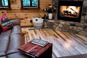 A cozy living room with a leather sofa, a small wooden table with a book, a stone fireplace, and a window with a view of trees. - Brand New Cabin | Theater | Game Room | Hot Tub (Logan)