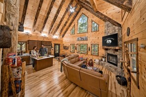 A log cabin interior with a stone fireplace, wooden beams, and a large window overlooking greenery. - Brand New Cabin | Theater | Game Room | Hot Tub (Logan)