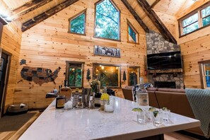 A wooden interior with a stone fireplace, a television, and a table with various bottles and glasses. - Brand New Cabin | Theater | Game Room | Hot Tub (Logan)