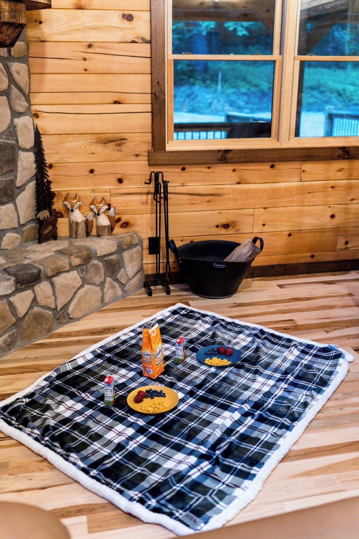 A cozy cabin interior with a plaid blanket on the floor, a stone fireplace, wooden walls, and a window with a view of trees.