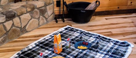 A cozy cabin interior with a plaid blanket on the floor, a stone fireplace, wooden walls, and a window with a view of trees.