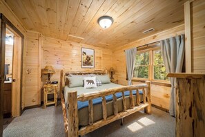 A wooden cabin room with a bed, a window with curtains, a lamp, and a picture on the wall.