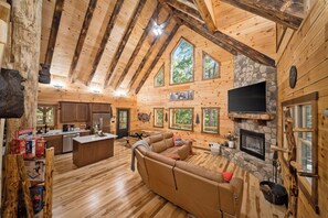 A spacious living area with a stone fireplace, wooden beams, and a large window overlooking greenery. - Brand New Cabin | Theater | Game Room | Hot Tub (Logan)