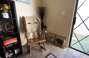 Une pièce d’angle avec un fauteuil à bascule en bois, une bibliothèque remplie de jeux, un vase de fleurs séchées et une fenêtre donnant sur un espace verdoyant.