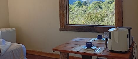 A room with a bed, a wooden table with a cup, a kettle, and a view of mountains.