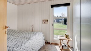 A bedroom with a wooden floor, a bed with a patterned bedspread, a small table with a box, and a door leading outside.