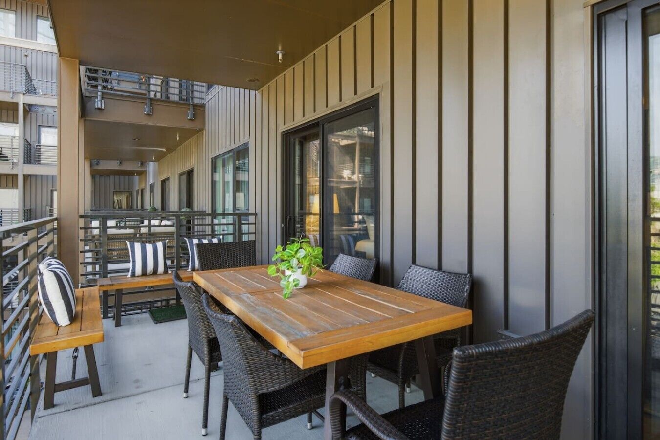 Un espace de restauration en plein air avec une table en bois et des chaises en osier, une plante en pot et une vue sur la ville à travers de grandes fenêtres.