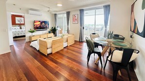 Een moderne woonkamer met een bank, eettafel en stoelen. Een televisie hangt aan de muur en er is een raam met gordijnen.