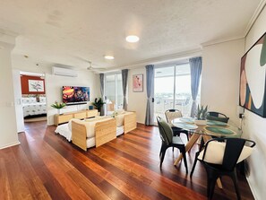 Ein modernes Wohnzimmer mit einer Couch, einem Esstisch und Stühlen. Ein Fernseher ist an der Wand montiert und es gibt ein Fenster mit Vorhängen.