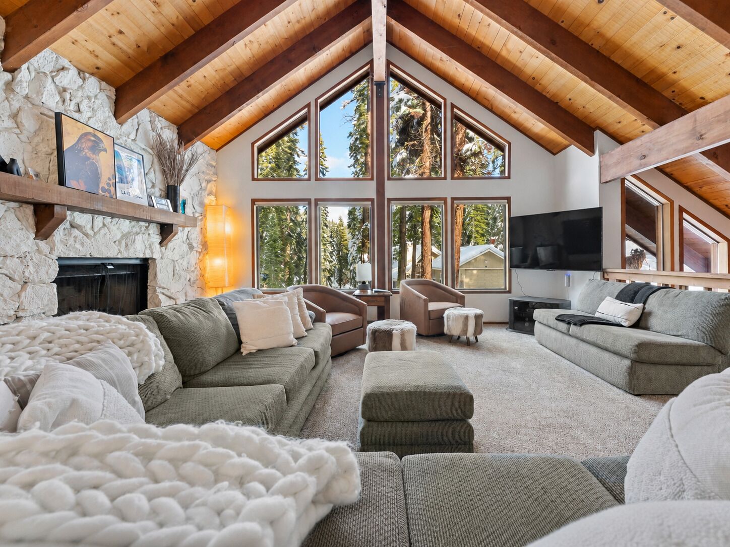 Ein geräumiges Wohnzimmer mit hohen Decken, einer Steinwand und großen Fenstern mit Blick auf Bäume.