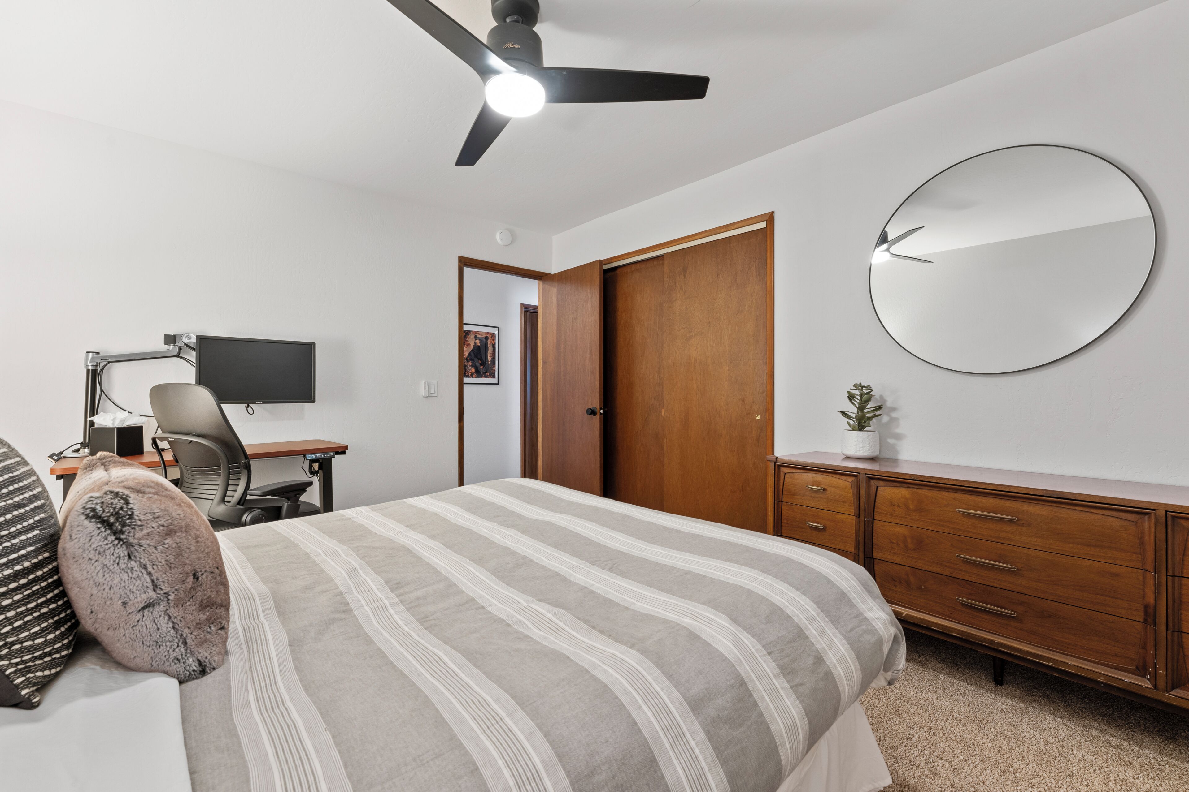 A bedroom with a bed, a desk, a chair, a computer, a ceiling fan, a wardrobe, a mirror, and a small plant.