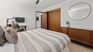 Un dormitorio con cama, escritorio, silla, computadora, ventilador de techo, armario, espejo y una planta pequeña.