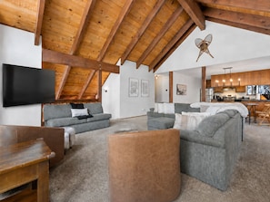 Ein geräumiger Wohnbereich mit hoher Holzbalkendecke, grauen Sofas, einem Flachbildfernseher und einer Küche im Hintergrund.