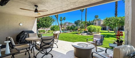 A patio area with a ceiling fan, outdoor furniture, and a view of a landscaped garden with palm trees and a pool.