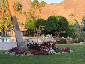 A pool area with lounge chairs, surrounded by a variety of flowers and palm trees, with mountains in the background. - Beautiful modern 2-bedroom villa with gorgeous views in Ironwood CC Palm Desert (Palm Desert)