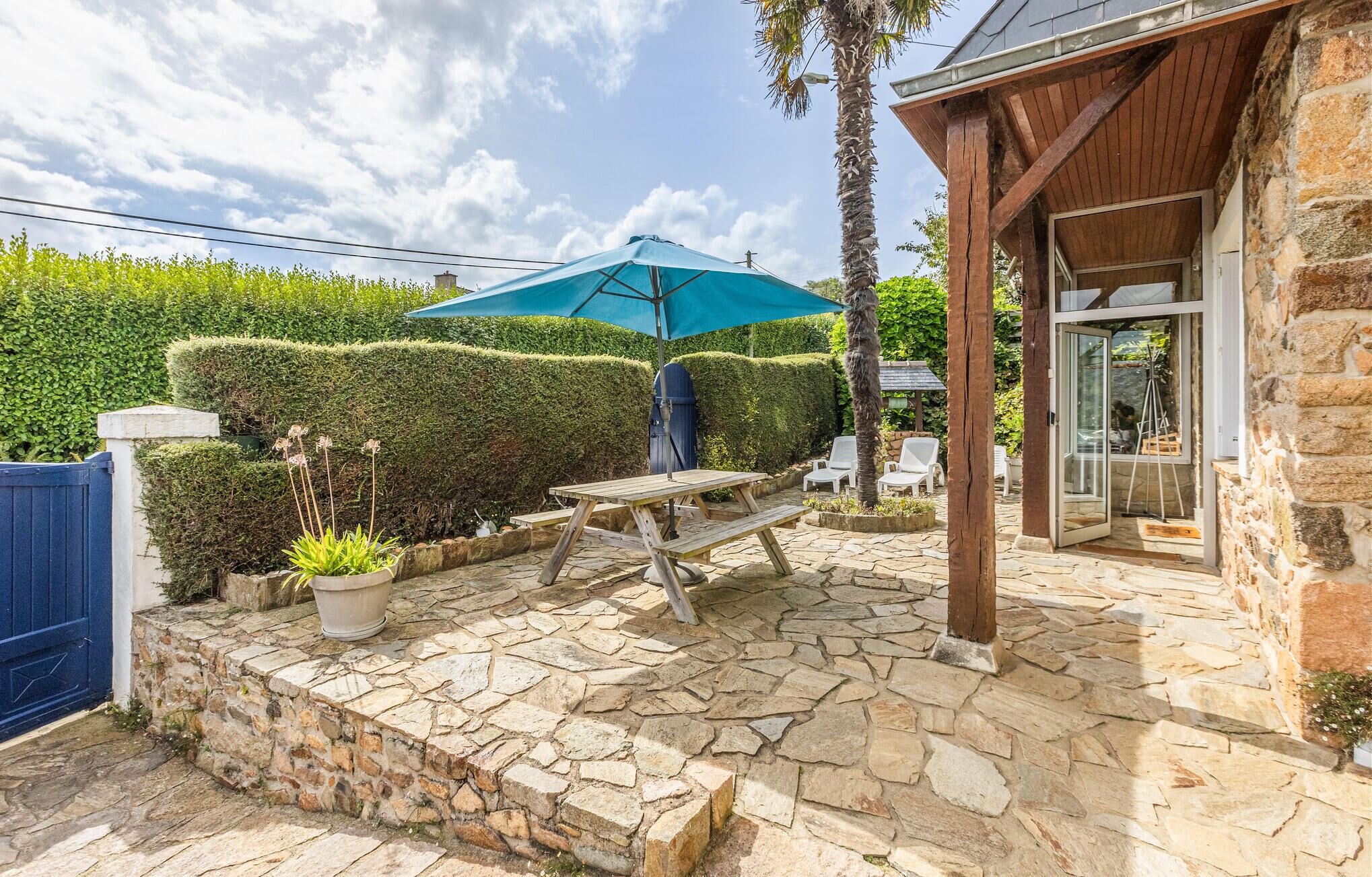 A stone patio with a wooden table and chairs, a blue umbrella, and a stone wall.