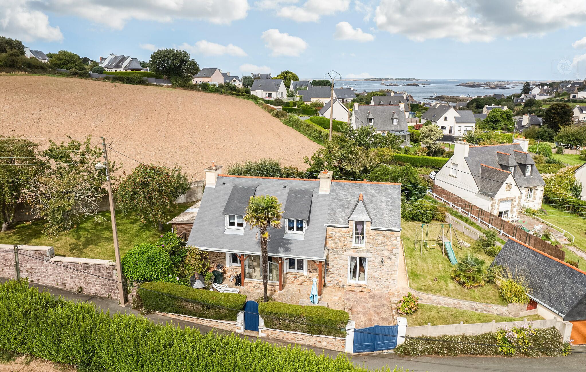 Una casa de piedra con techo inclinado, rodeada de exuberante vegetación y vista al mar.