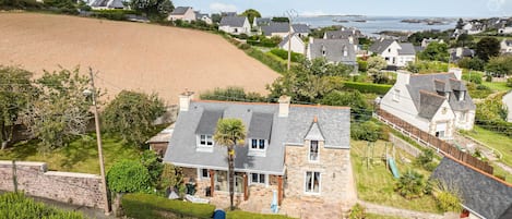 Una casa de piedra con techo inclinado, rodeada de exuberante vegetación y vista al mar.