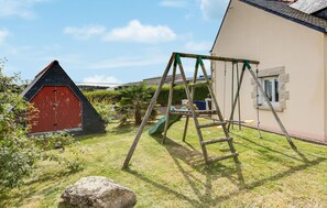 A wooden swing set in a grassy yard with a red barn and a house in the background. - Amazing home in Ploubazlanec with WiFi (Ploubazlanec)