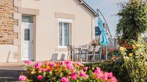 Een gebouw met een gevel van steen, een deur en ramen. Er is een terras met stoelen en een tafel, en een blauwe parasol.