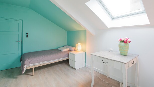 Een slaapkamer met een schuine kap, dakraam, een bed, nachtkastje, een dressoir en een vaas met bloemen.