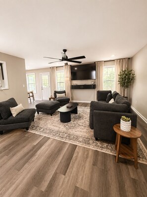 A modern living room with a flat-screen TV, dark sofas, a coffee table, and a wooden floor. - The Key Manor Retreat Modern Stay Near Atlanta (Lawrenceville)