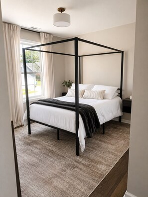 Ein Bett mit Baldachin in Weiß und Schwarz, ein Nachttisch mit Lampe und Blick ins Freie durch ein Fenster.