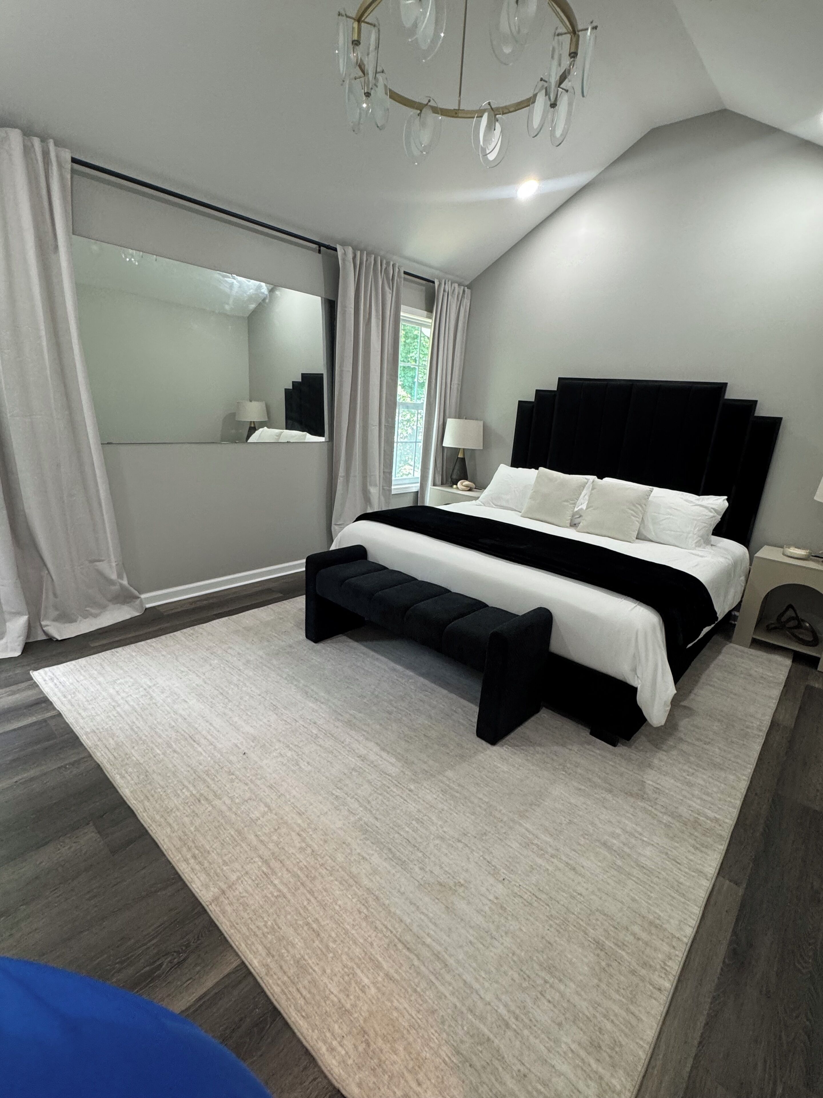 A modern bedroom with a large bed, a bench, a nightstand, a mirror, and a chandelier.