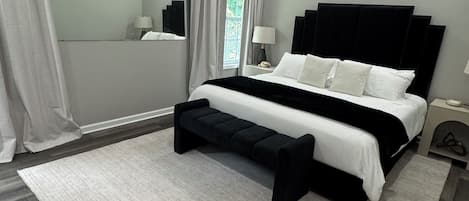 A modern bedroom with a large bed, a bench, a nightstand, a mirror, and a chandelier.