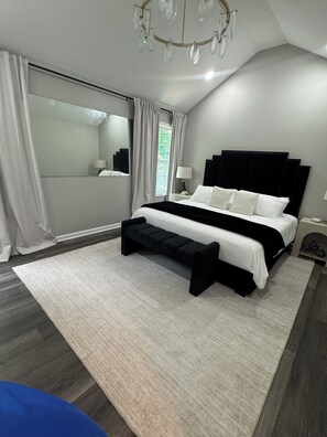 Un dormitorio moderno con una cama grande, un banco, una mesita de noche, un espejo y una araña.