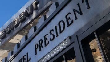 Um edifício com uma placa indicando 'HOTEL PRESIDENT EST. 1928 - LUXURY ACCOMMODATIONS' e uma construção alta ao fundo.