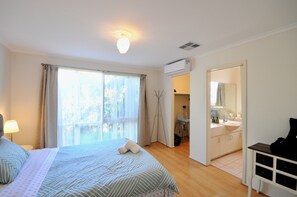 A hotel room with a bed, a window with curtains, a bedside lamp, a bathroom with a sink and mirror, and an air conditioning unit. - Comfy unit on a quiet low traffic street. Brand new kitchen, central in Melb S/E (Glen Waverley)