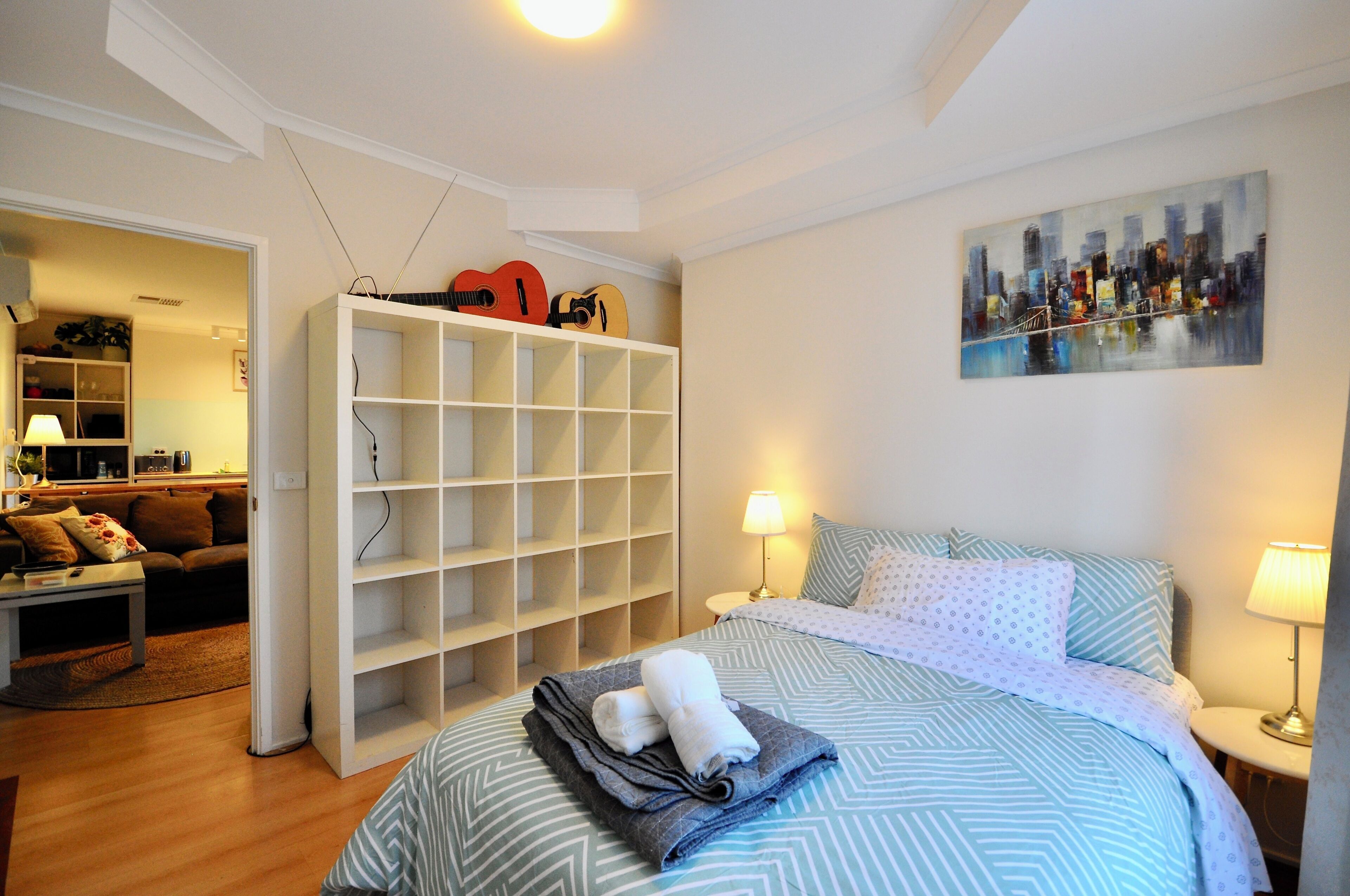 Una camera da letto con un letto, due abat-jour, un dipinto con vista sulla città, un armadio bianco e un tavolino con un telecomando.