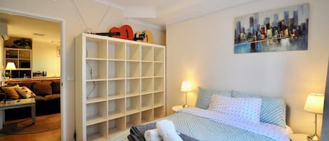 Un dormitorio con cama, dos lámparas de noche, un cuadro de paisaje urbano, un armario blanco y una pequeña mesa con un control remoto.