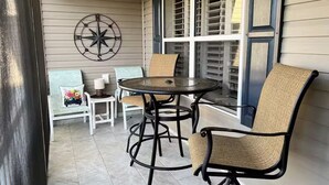 Une terrasse couverte avec une table ronde et des chaises, une décoration murale en forme de boussole et une fenêtre avec des volets.