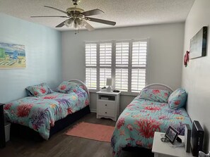Ein Schlafzimmer mit zwei Betten, einem Deckenventilator, einem Fenster mit Jalousien, einem Nachttisch und einem Schreibtisch mit Computer.