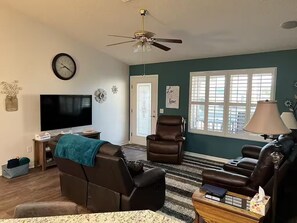 Ein Wohnzimmer mit Flachbildfernseher, Ledersofas, Deckenventilator und einem Fenster mit Jalousien.