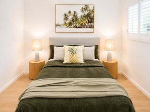 A bedroom with a bed, two bedside lamps, a framed picture on the wall, and a window with blinds.