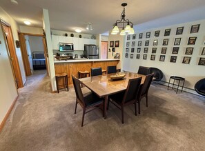 A dining area with a wooden table and chairs, a kitchen in the background, and a wall with framed pictures.