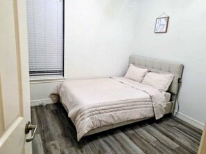 Ein Schlafzimmer mit Bett, einem Fenster mit Jalousien und einem an der Wand hängenden Bilderrahmen.