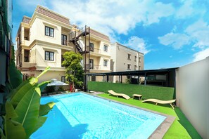 Una piscina con acqua cristallina, circondata da un'area di prato sintetico verde, adiacente a un edificio a più piani con balconi.