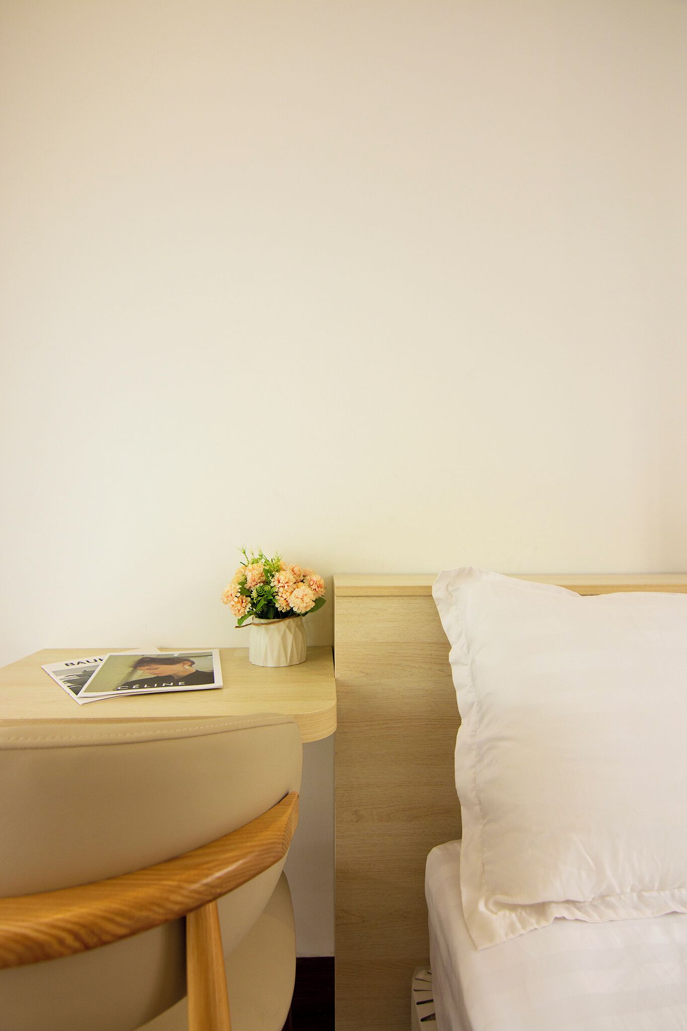 Una habitación de hotel con cama, mesita de noche, una silla y una pequeña mesa con una revista y un florero.