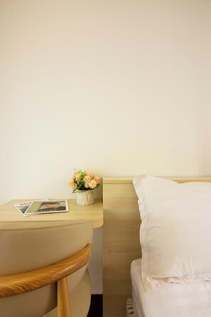 Una habitación de hotel con cama, mesita de noche, una silla y una pequeña mesa con una revista y un florero.