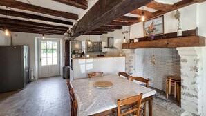 A rustic kitchen with exposed wooden beams, a dining table with chairs, and a fireplace. - Nice home in Manthelan (Manthelan)
