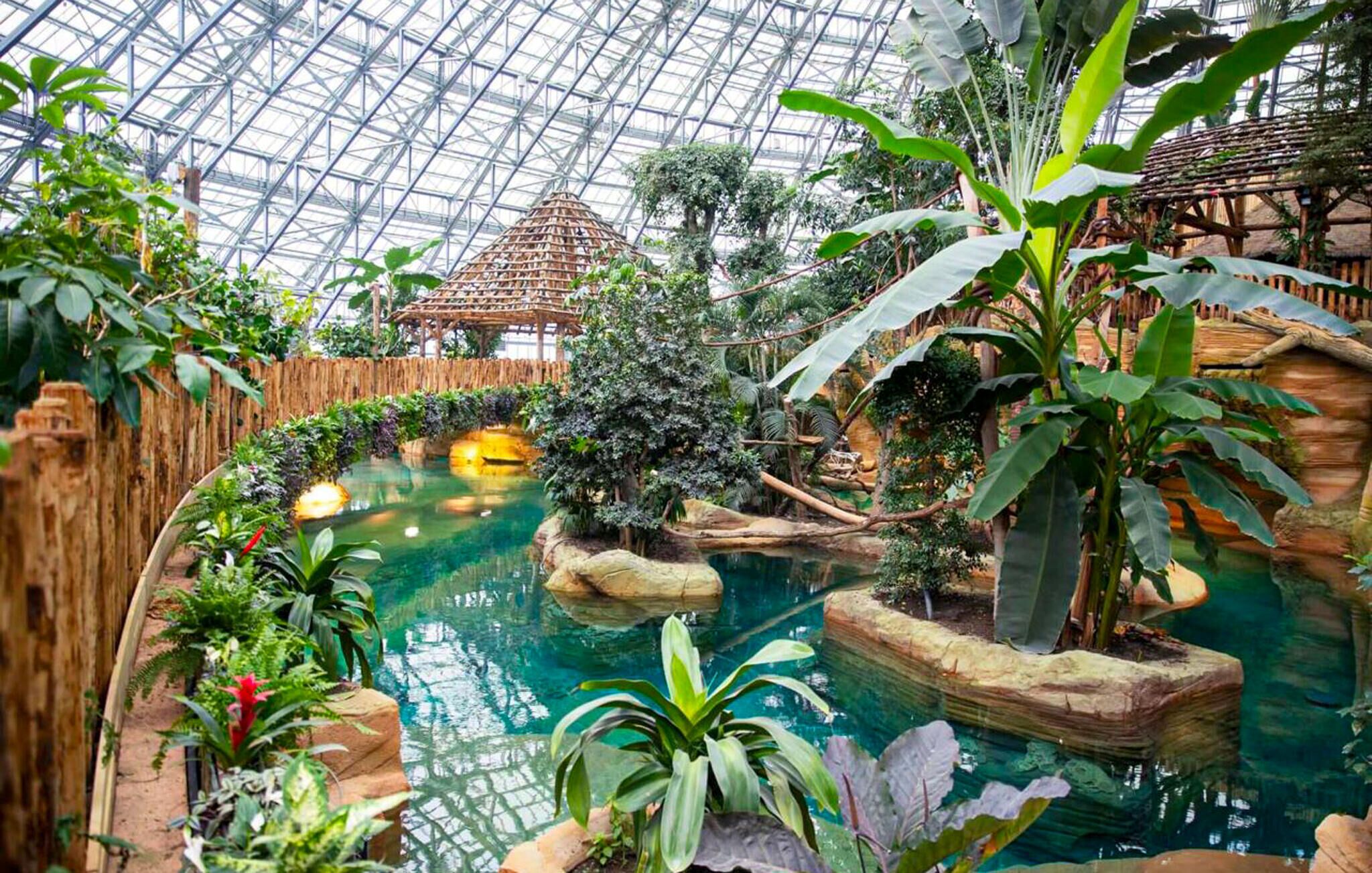 Ein botanischer Garten mit Glasdach, Wasserspiel und verschiedenen tropischen Pflanzen.